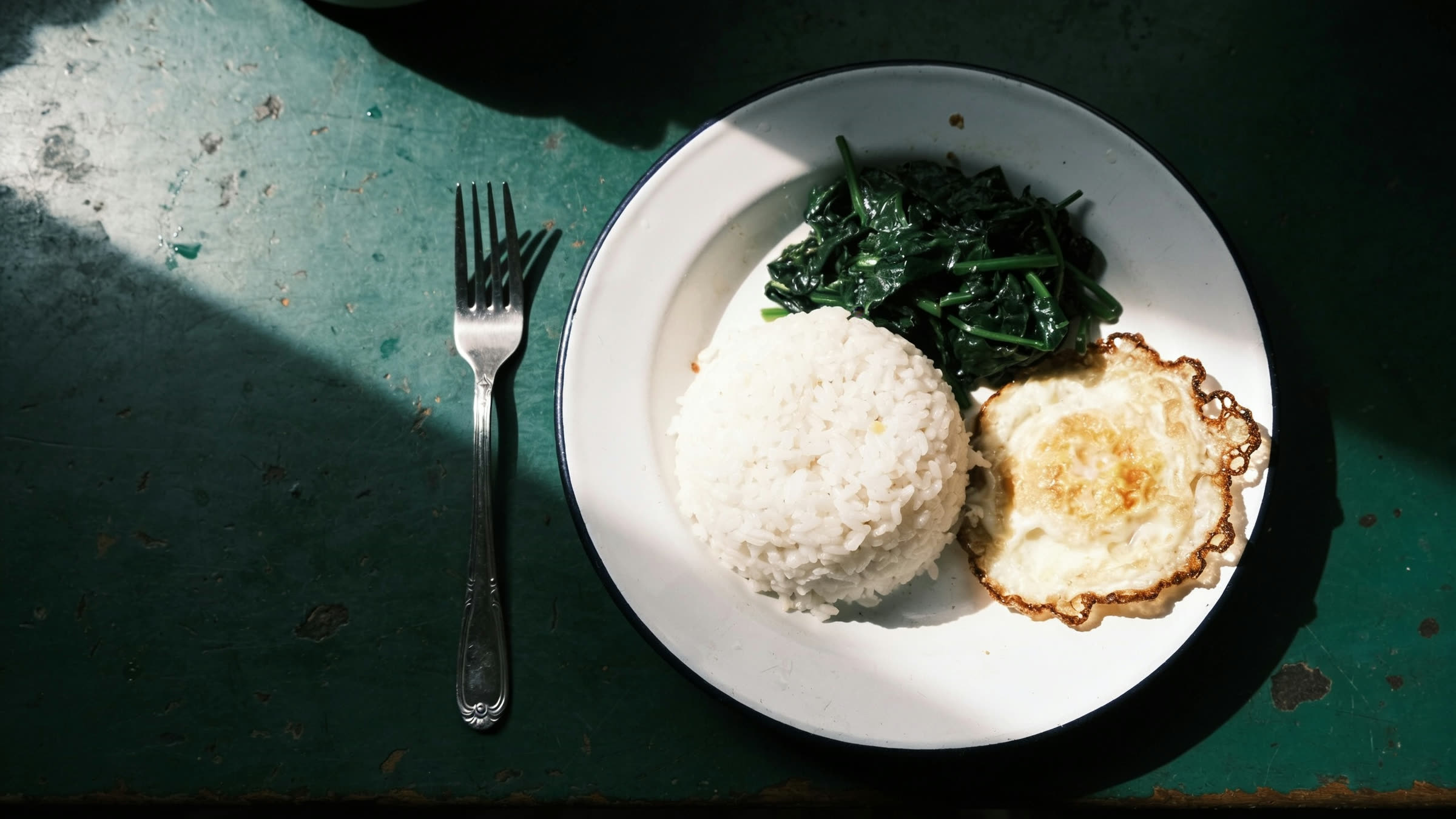 Rice and greens with a fried egg on a plain plate, photographed flat under one hard light.